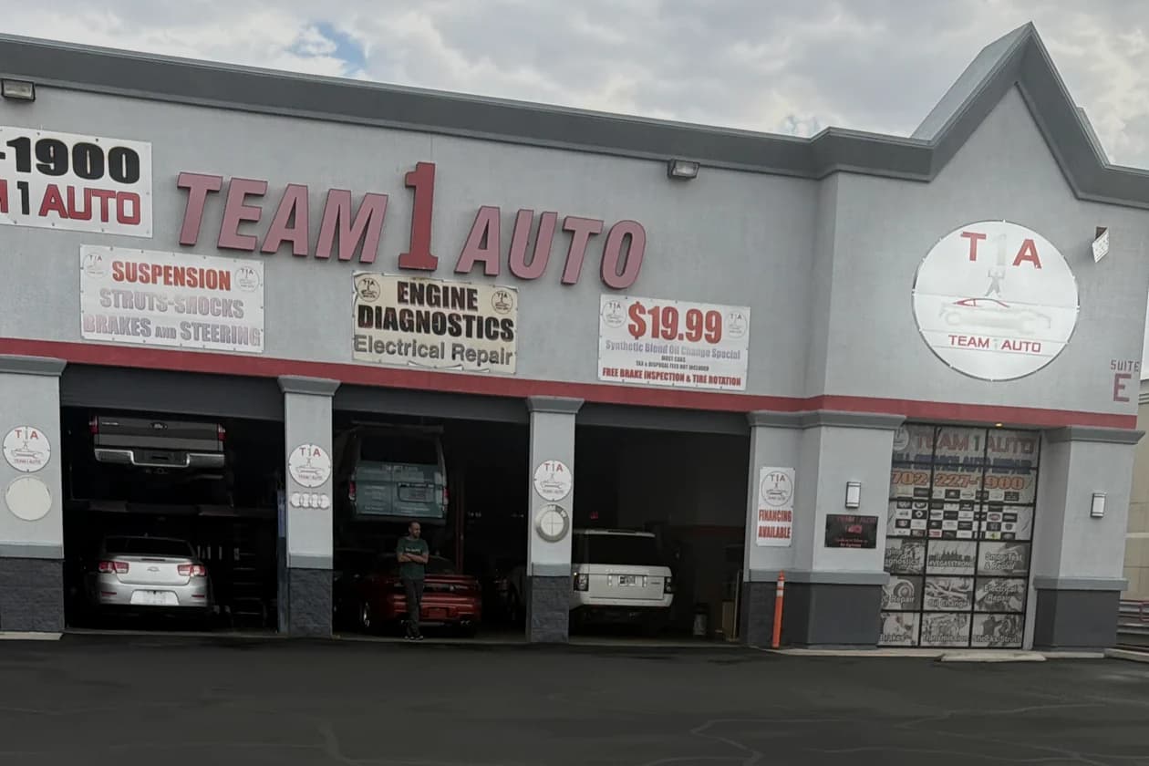 The Team 1 Auto Care building on W Russell Rd: three open service bays, red TEAM 1 AUTO lettering, and a white circular T1A sign on the peaked gable above Suite E.